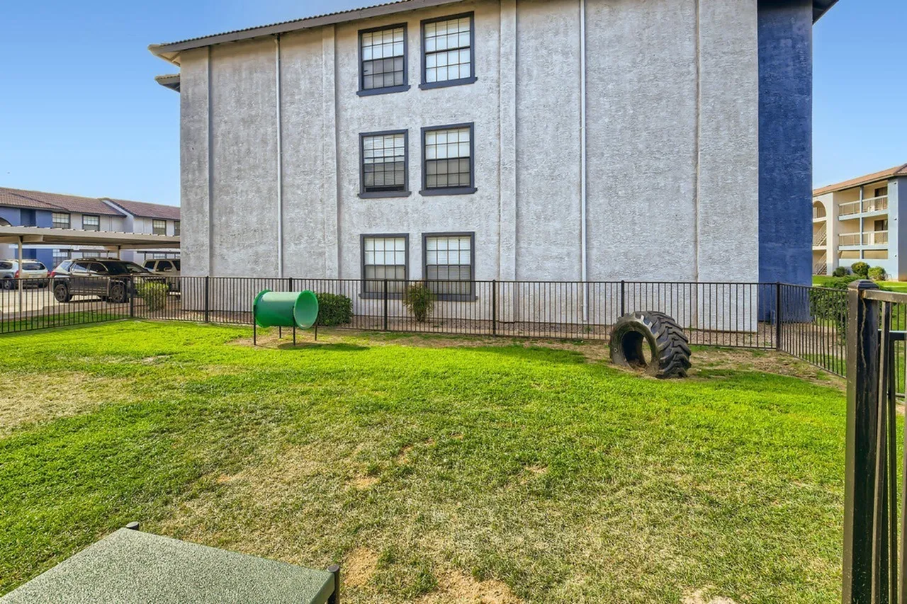 Grassy courtyard with playground equipment, metal fencing, three-story residential building