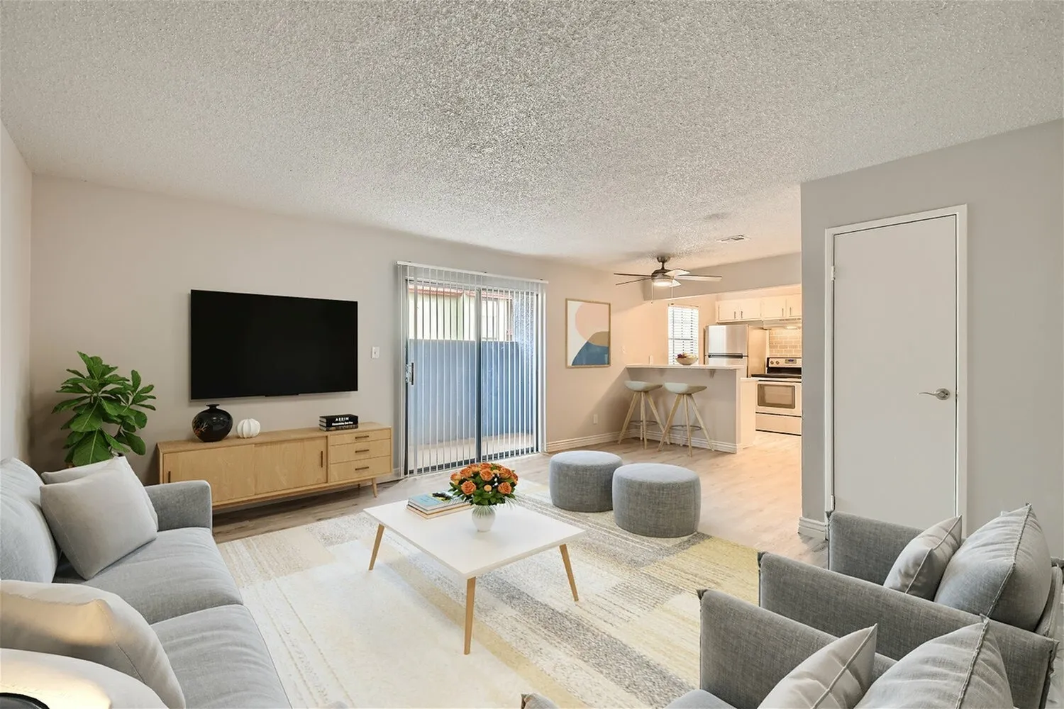 Living room with wood-style plank flooring, gray upholstered seating, wall-mounted TV, open kitchen with light cabinetry and breakfast bar