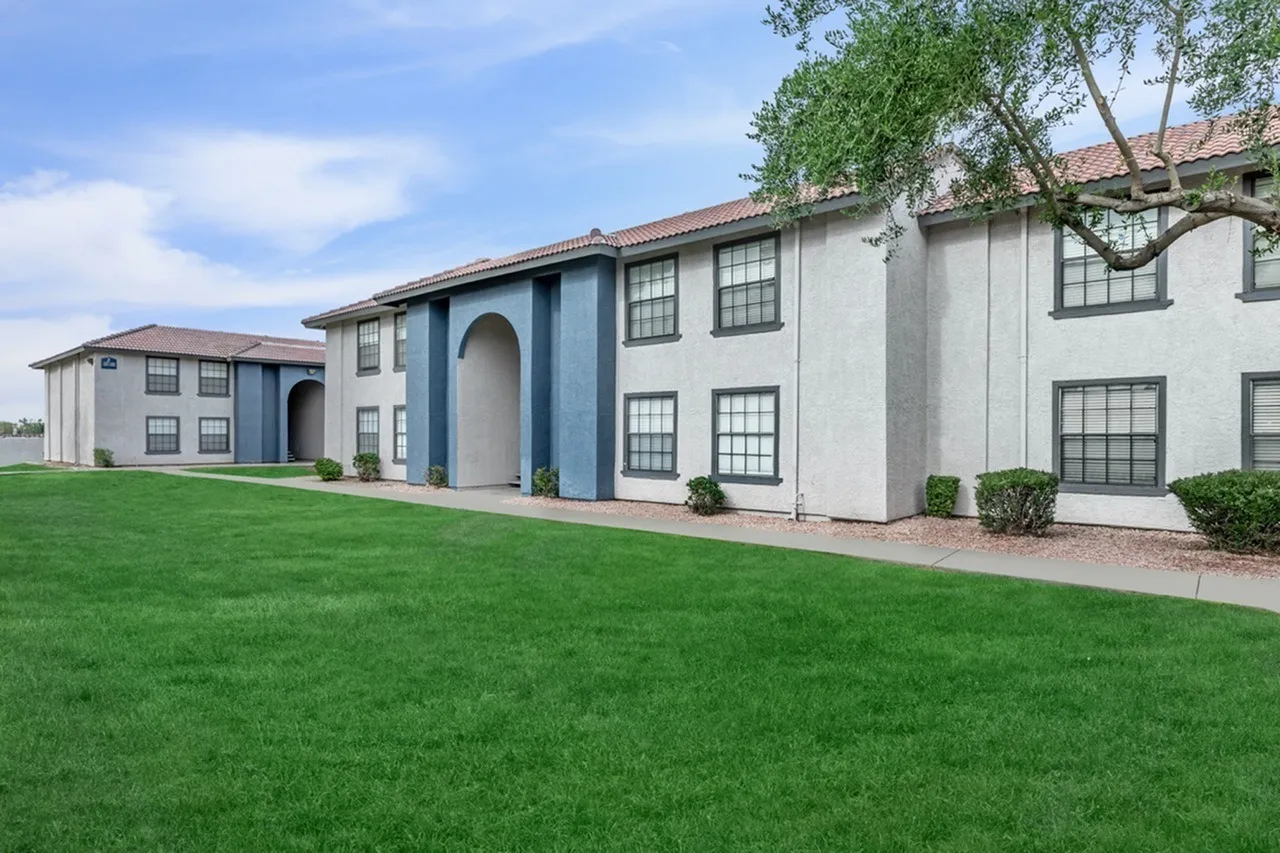 Residential building with white and blue stucco facade, arched entryway, multiple windows, clay tile roofing, manicured lawn