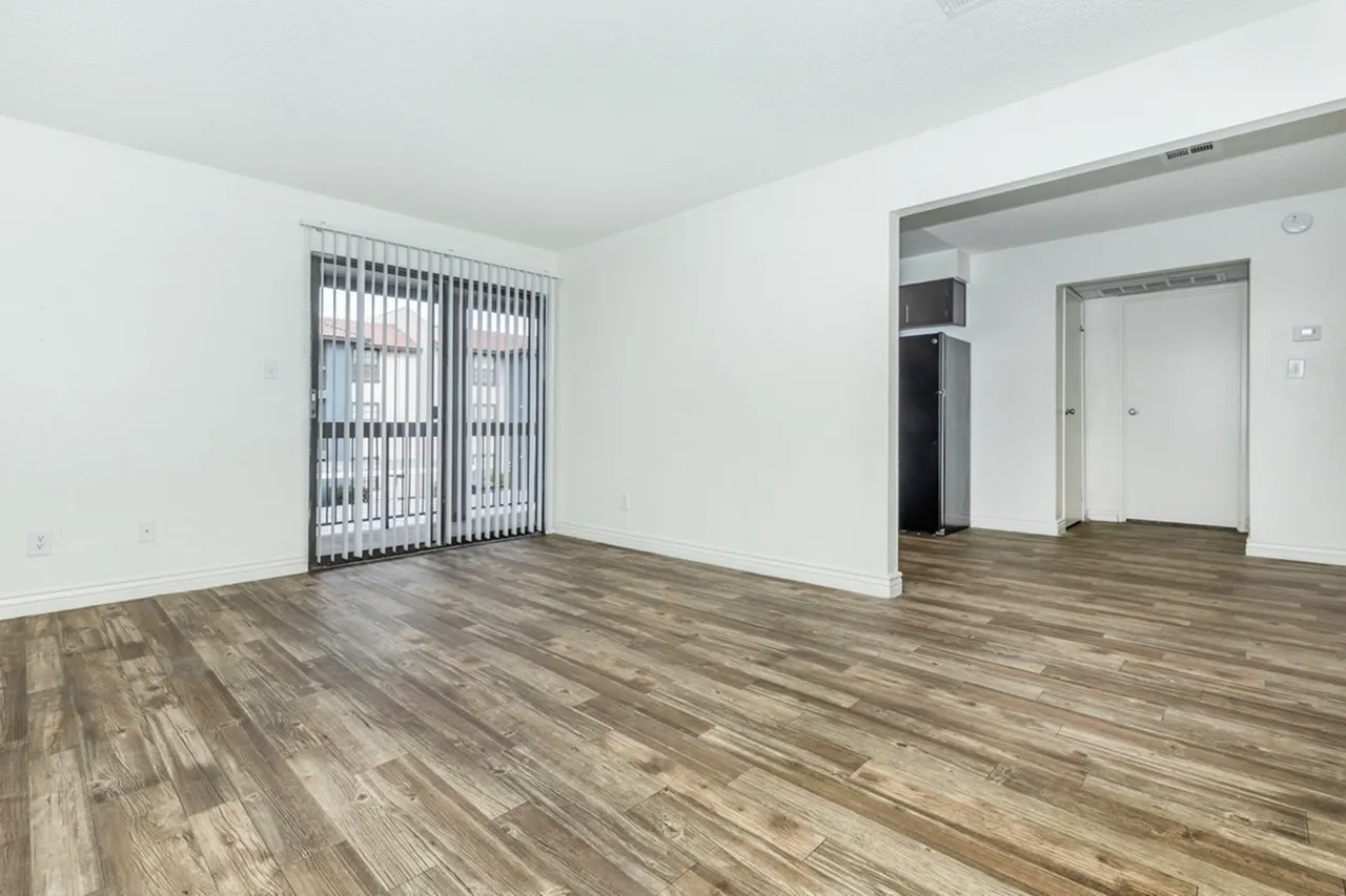 Living room with wood-style plank flooring, vertical blinds, white walls, entry door