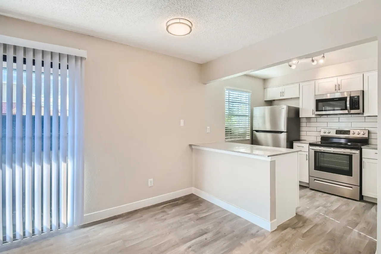 Kitchen with white shaker cabinetry, stainless steel appliances, white subway tile backsplash, wood-style plank flooring, and adjacent living space with vertical blinds
