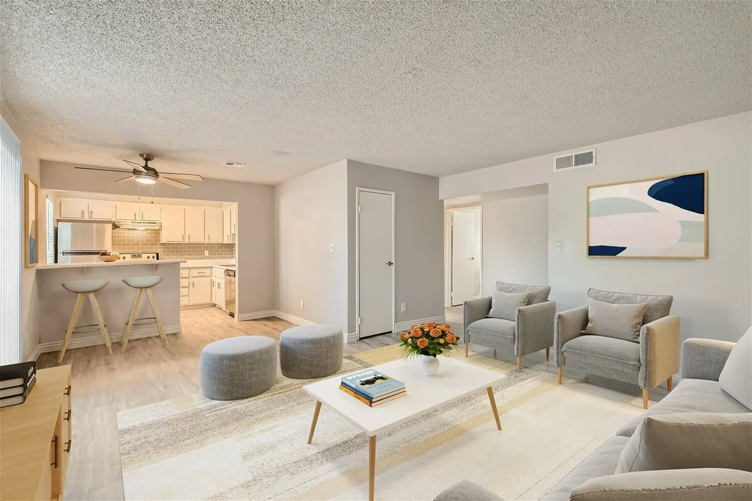 Living room with wood-style plank flooring, gray upholstered chairs, white coffee table, ceiling fan, kitchen with light cabinetry visible