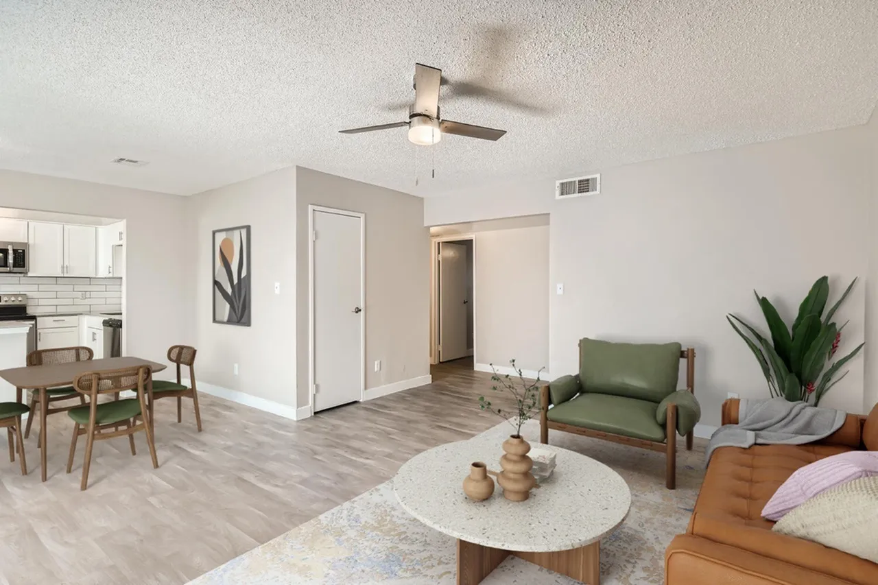 Living room with wood-style plank flooring, ceiling fan, green accent chair, round coffee table, dining area with wood table and chairs, kitchen visible