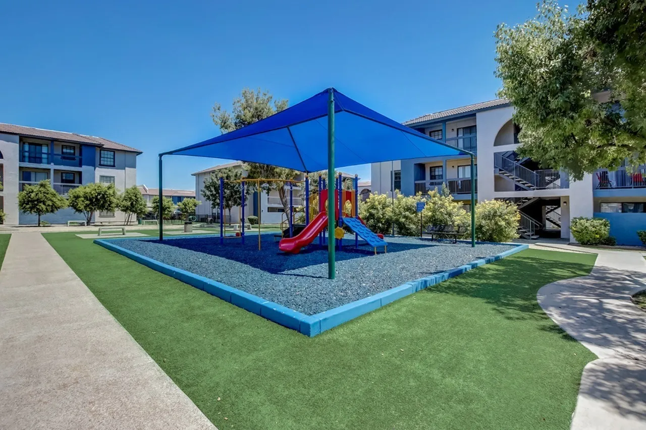 Playground with blue shade structure, red and blue slide, mulch surfacing, grass lawn, residential buildings