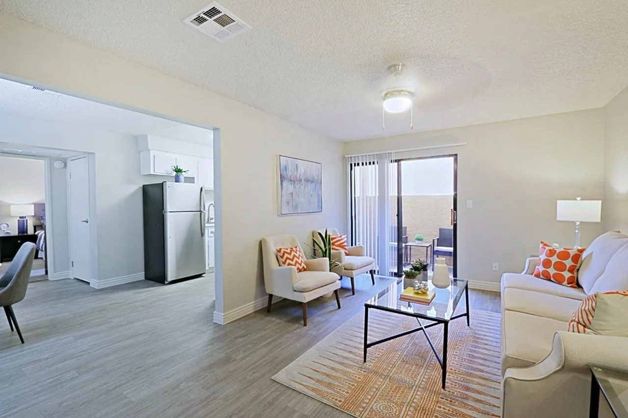 Living room with wood-style plank flooring, area rug, accent chairs with orange pillows, coffee table, ceiling lights, kitchenette with refrigerator, vertical blinds, glass doors to patio
