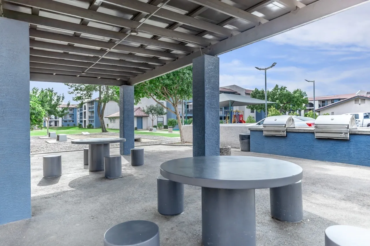 Outdoor covered pavilion, concrete round tables with seating stools, blue support columns, metal pergola roof, green landscape