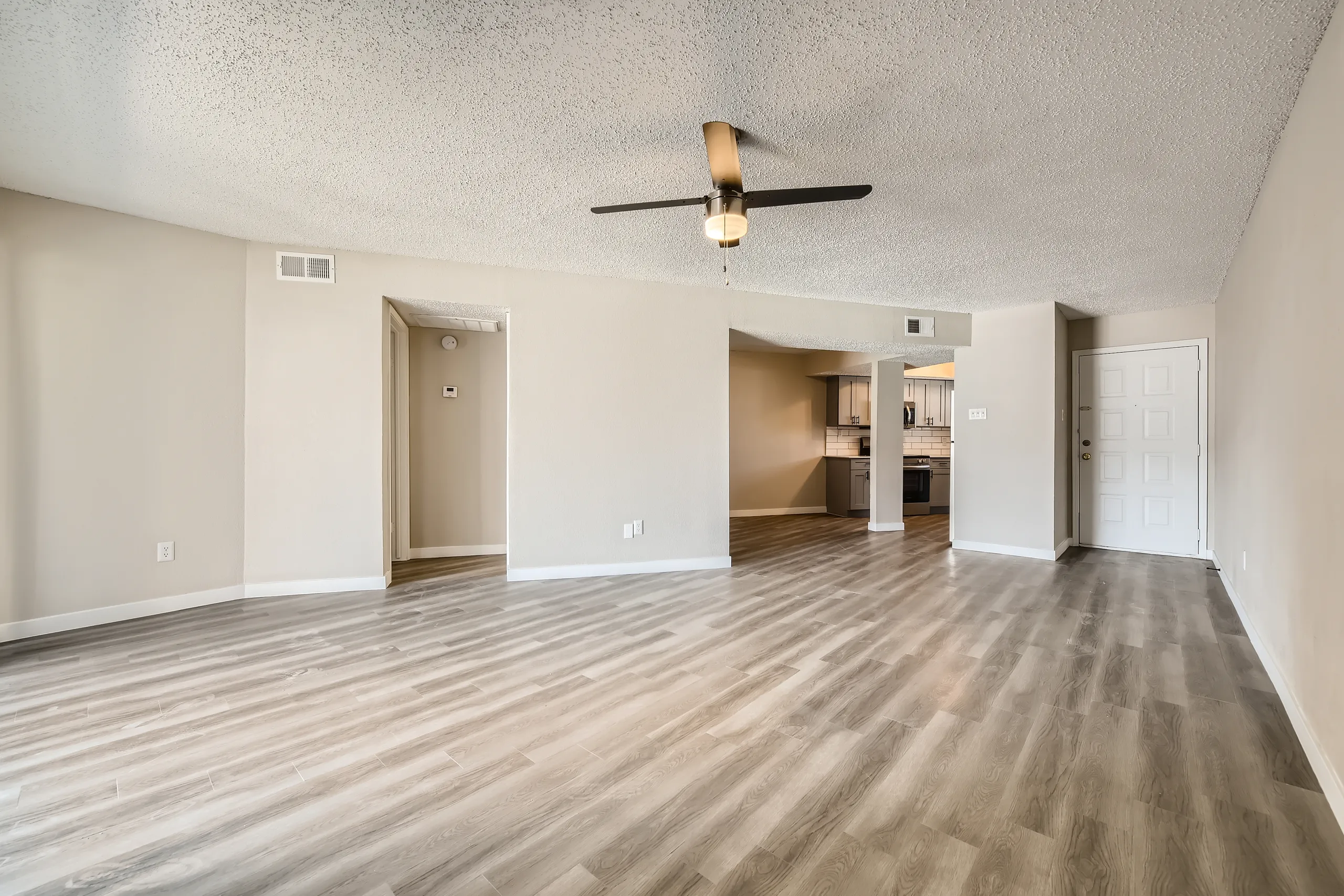 Open living room with wood-style plank flooring, ceiling fan, entry door, view into kitchen