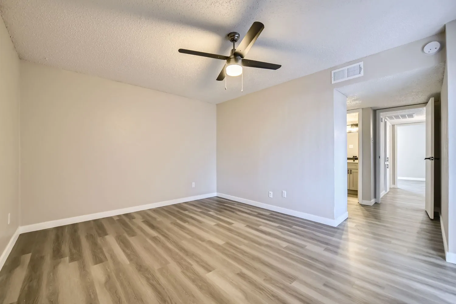 Primary bedroom with wood-style plank flooring, ceiling fan, hallway view to en-suite bathroom