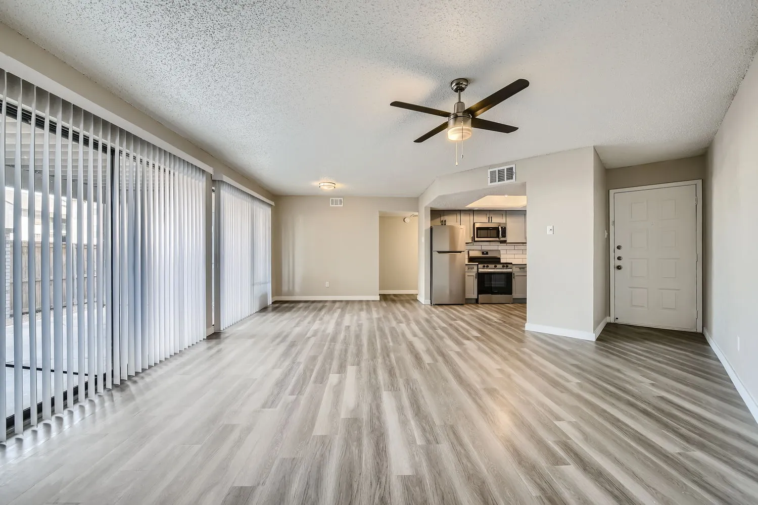 Living room with wood-style plank flooring, ceiling fan, entry door, view into kitchen