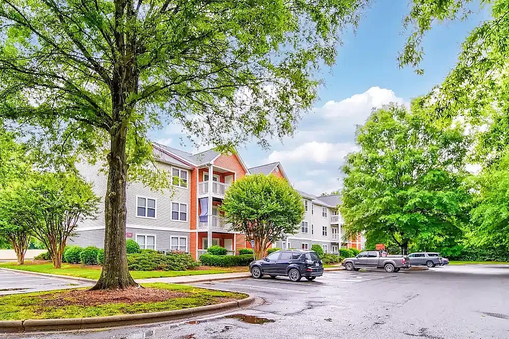 Apartment building exterior, red brick facade, mature trees, manicured landscaping, parked vehicles