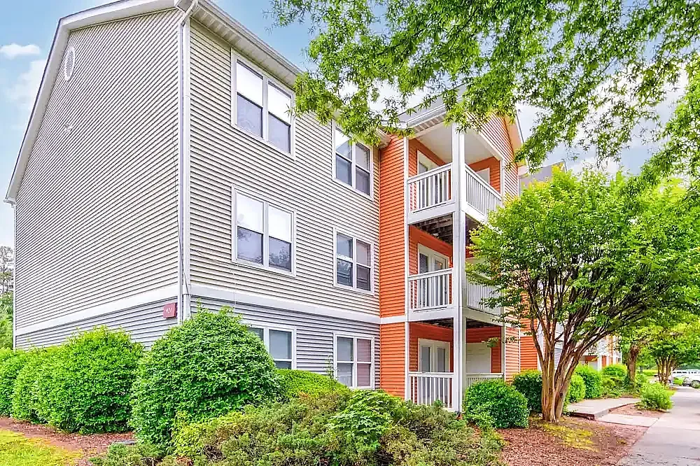 Multi-story apartment building with gray vinyl siding and orange brick accent, white railings, green landscaping and mature trees