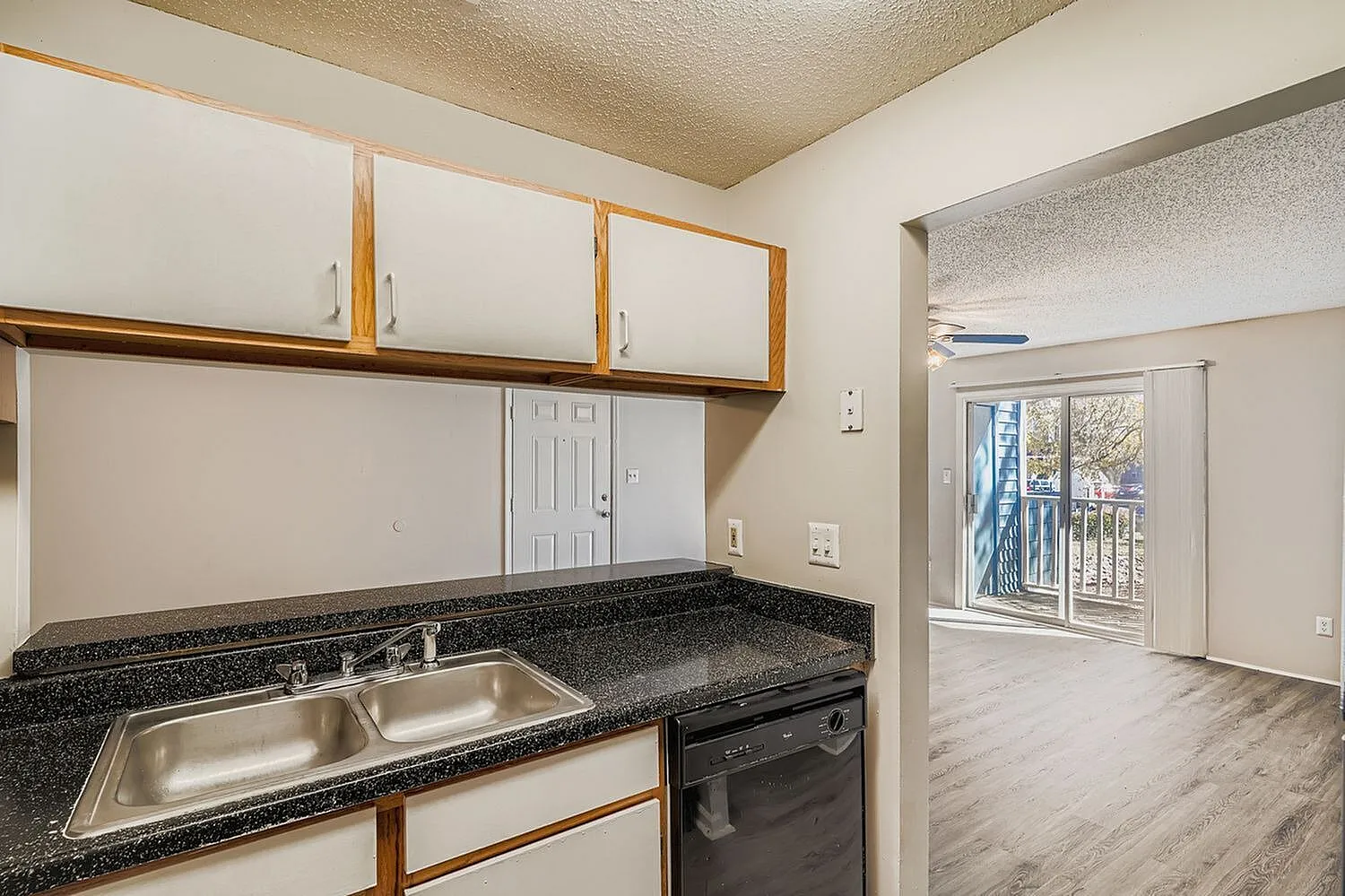 Kitchen with speckled black countertops, stainless steel sink, wood cabinetry, black appliances, view to living area with wood-style plank flooring and sliding glass doors