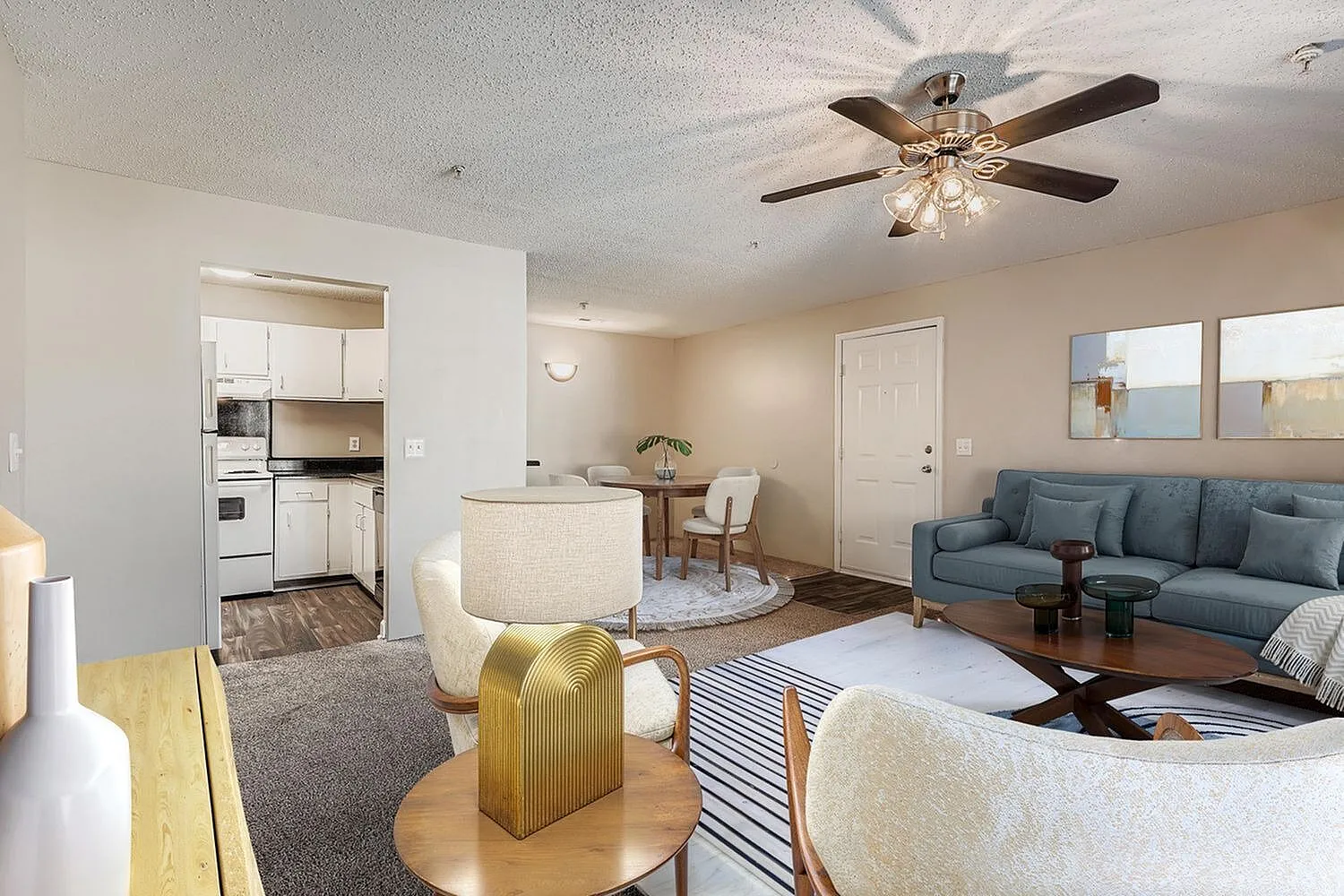 Open concept living area, wood-style plank flooring, cream upholstered chairs, blue sofa, ceiling fan