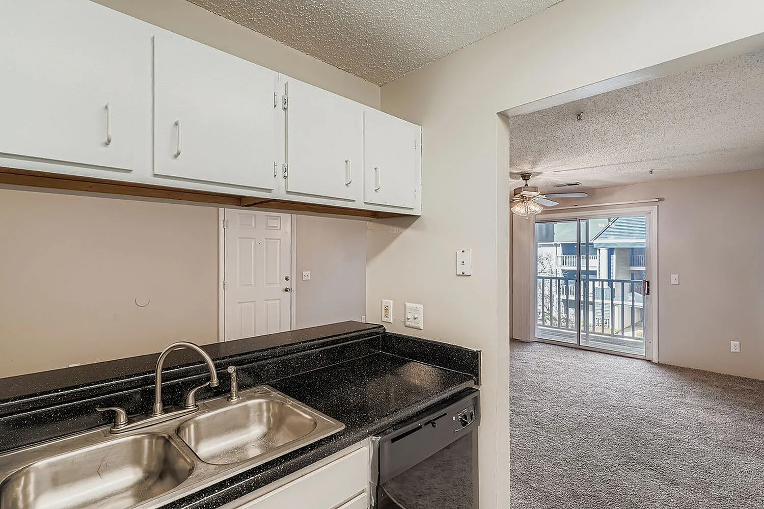 Kitchen with white shaker cabinetry, black granite countertops, stainless steel sink, connected to living area with wood-style plank flooring and glass sliding door