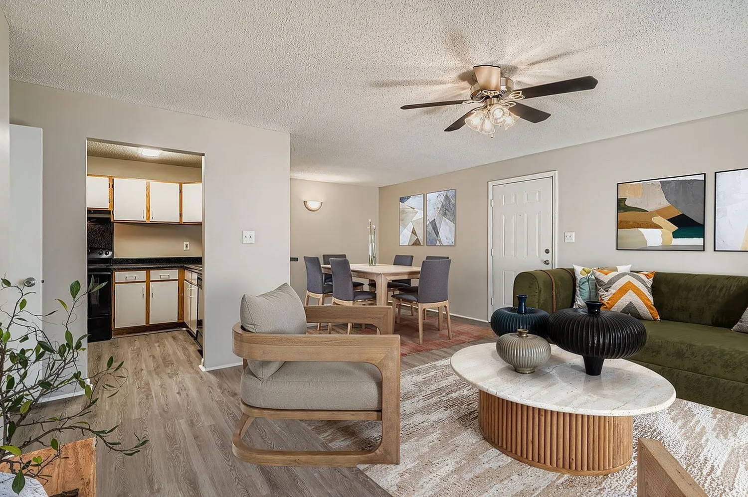 Living room with wood-style plank flooring, ceiling fan, green upholstered sofa, round wood coffee table, dining area, kitchen with wood cabinetry