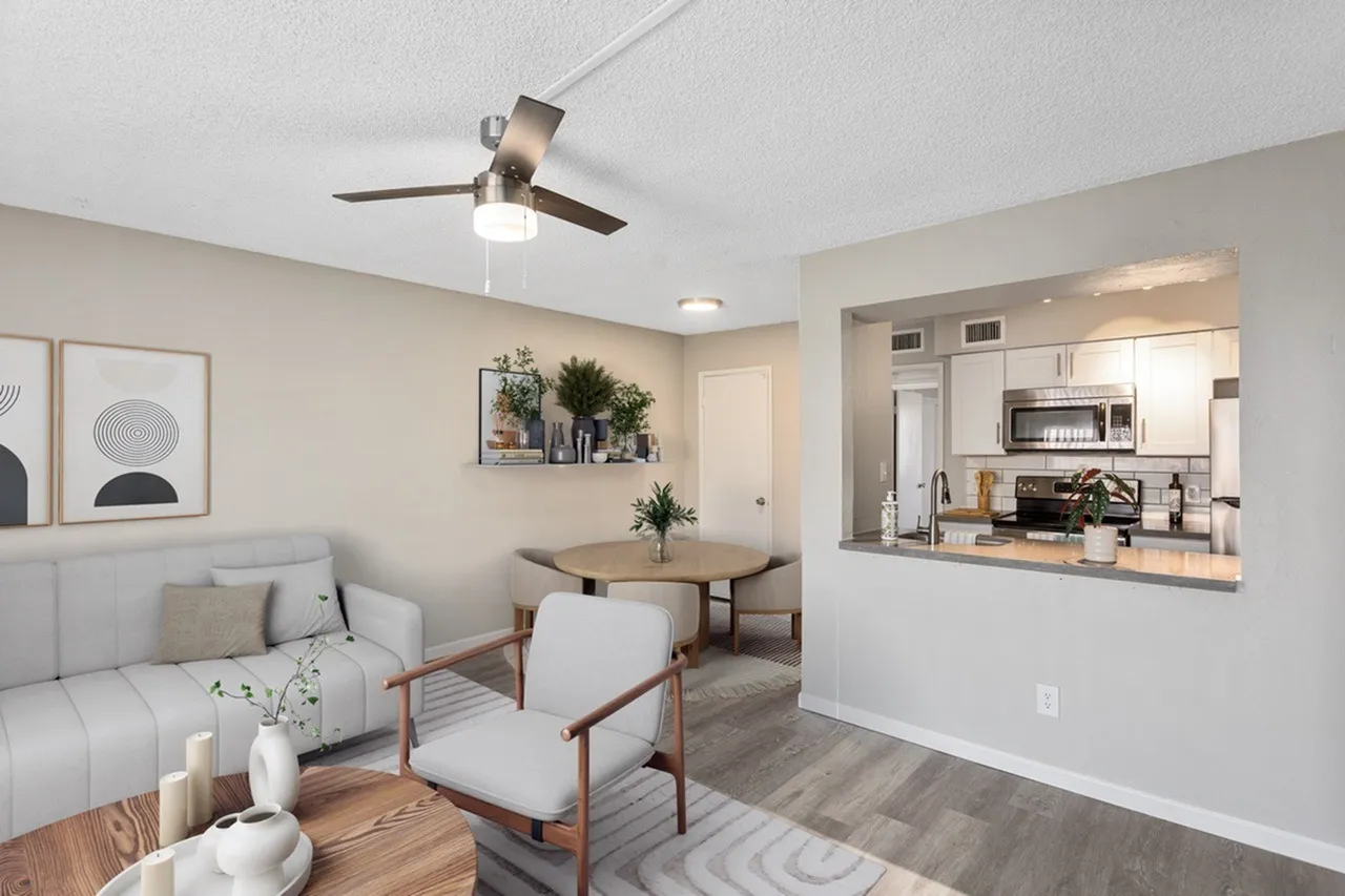 Living room with wood-style plank flooring, ceiling fan, beige sectional sofa, accent chair with wood frame, open to kitchen with white cabinetry and stainless appliances
