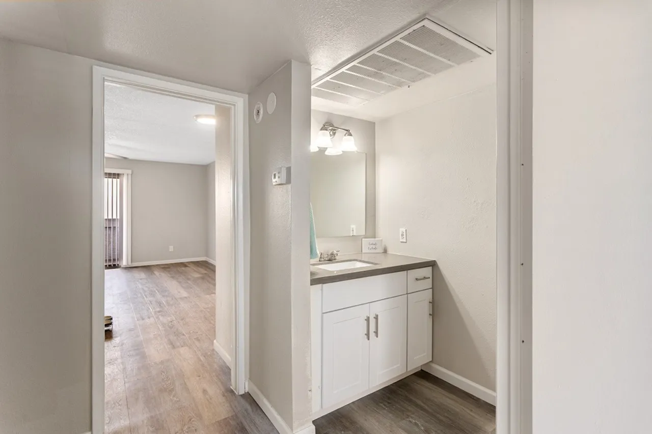 Bathroom vanity with white cabinetry and countertop, large mirror with lighting, hallway with wood-style plank flooring