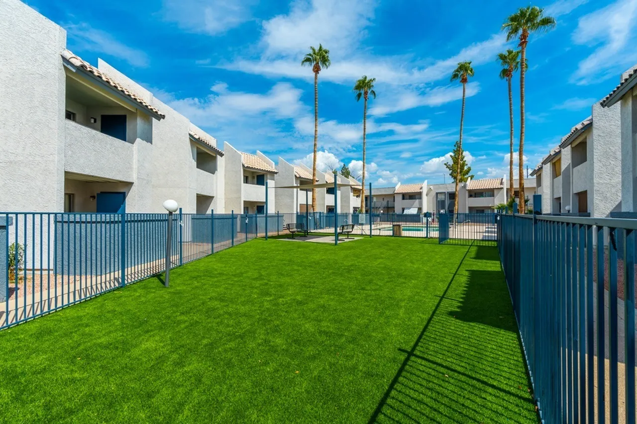 Central courtyard with green lawn, blue metal fencing, white residential buildings, palm trees, clear sky