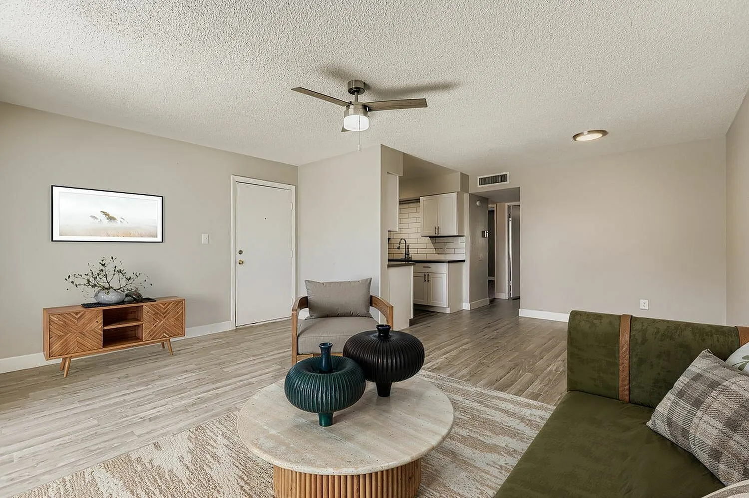 Living room with wood-style plank flooring, green upholstered sofa, ceiling fan, round coffee table with decorative vessels, mid-century wood sideboard with wall art above, kitchen visible through open layout