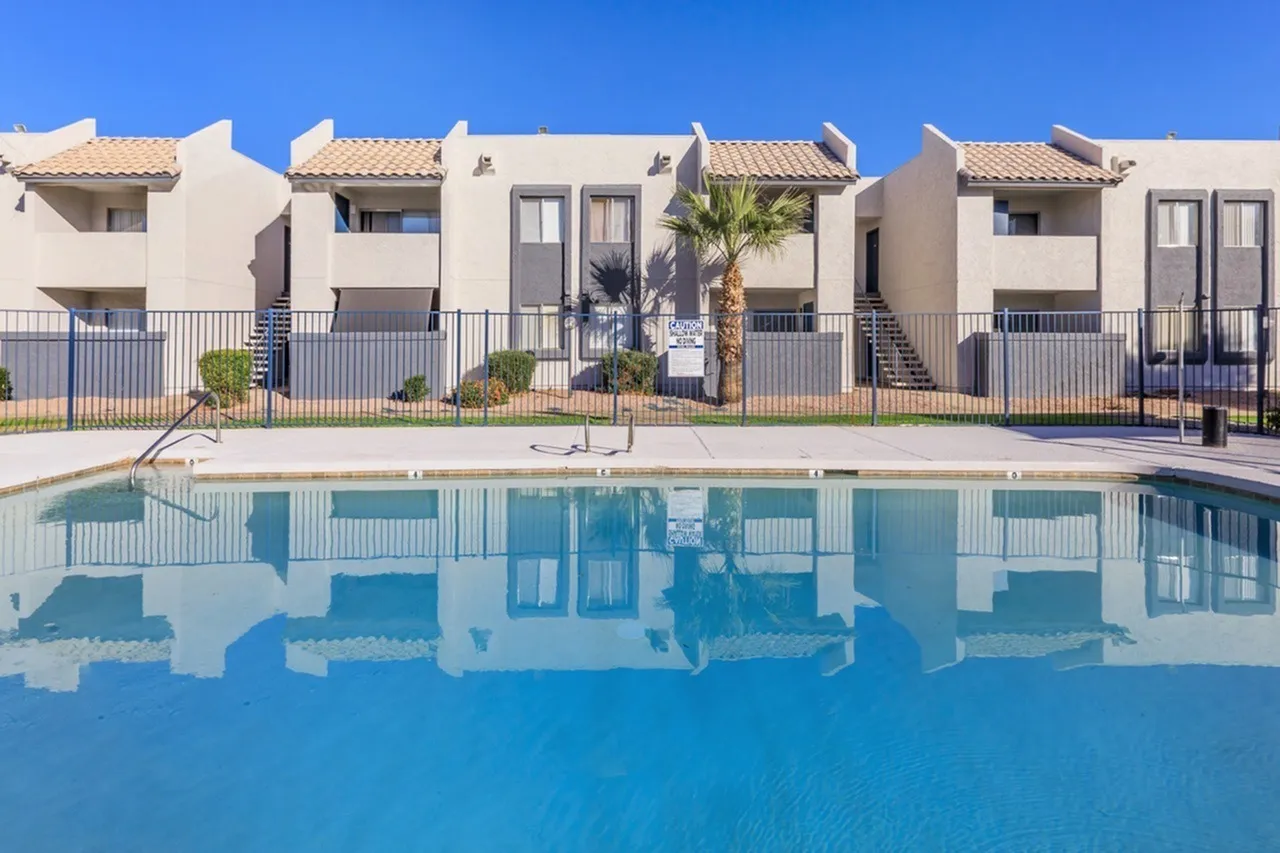 Community swimming pool with clear blue water, tan townhomes with tile roofs, metal fence, palm tree, clear sky