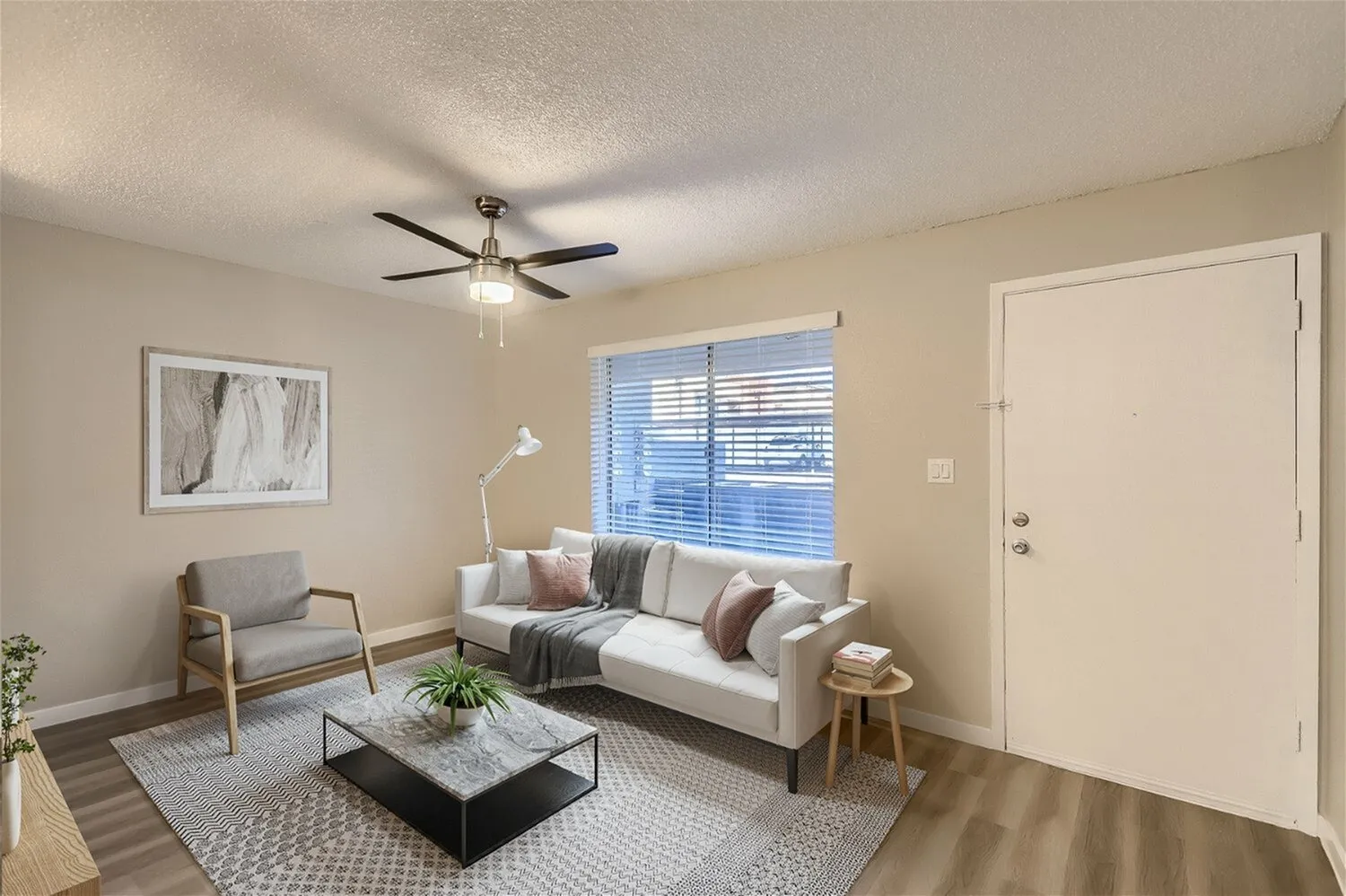 Living room, wood-style plank flooring, beige sofa with throw pillows, gray accent chair, ceiling fan, window with blinds
