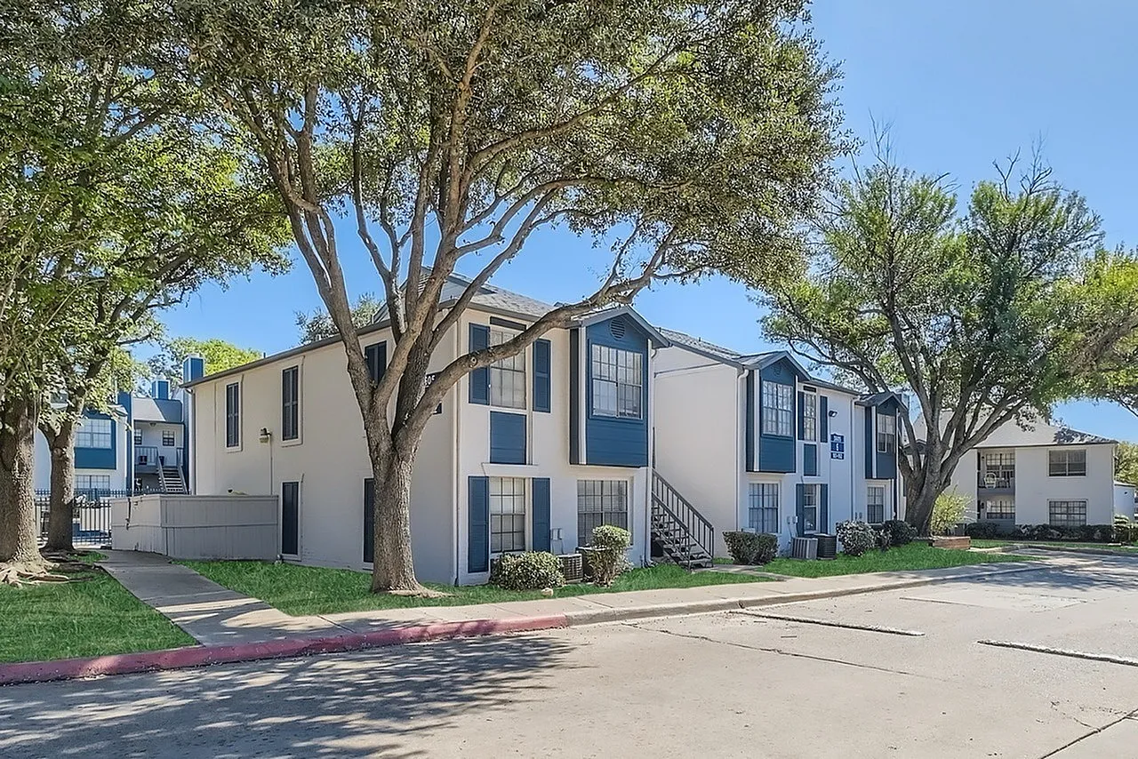 Multi-story apartment building, white and blue facade, mature trees, paved parking area
