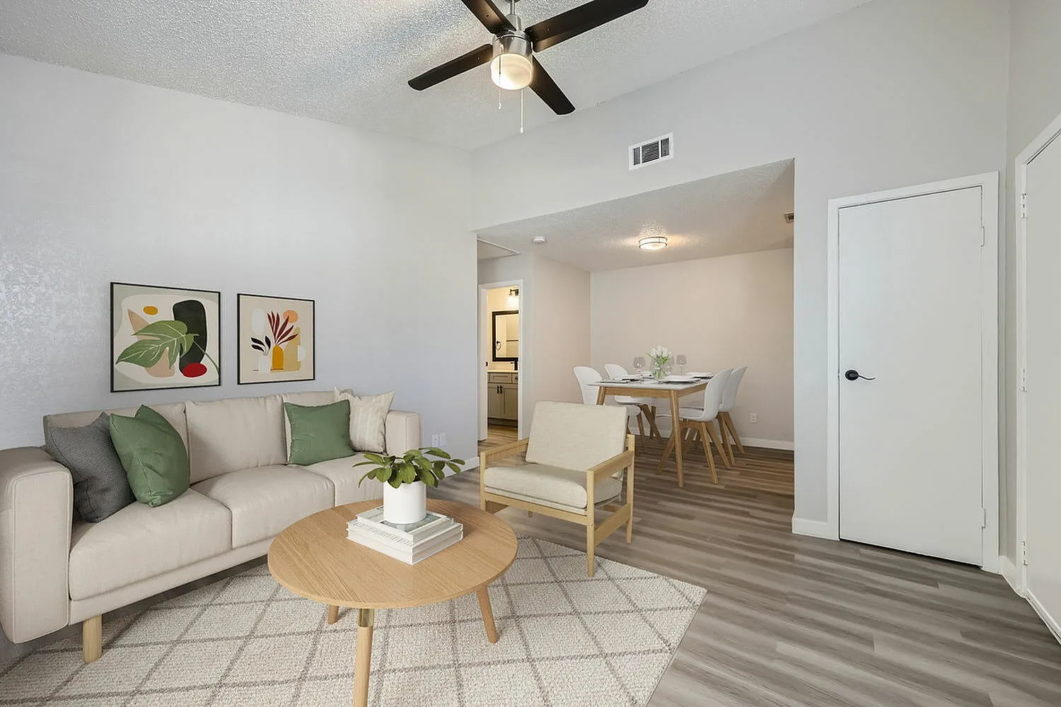 Living room with wood-style plank flooring, beige sofa with green pillows, ceiling fan, dining area with white chairs