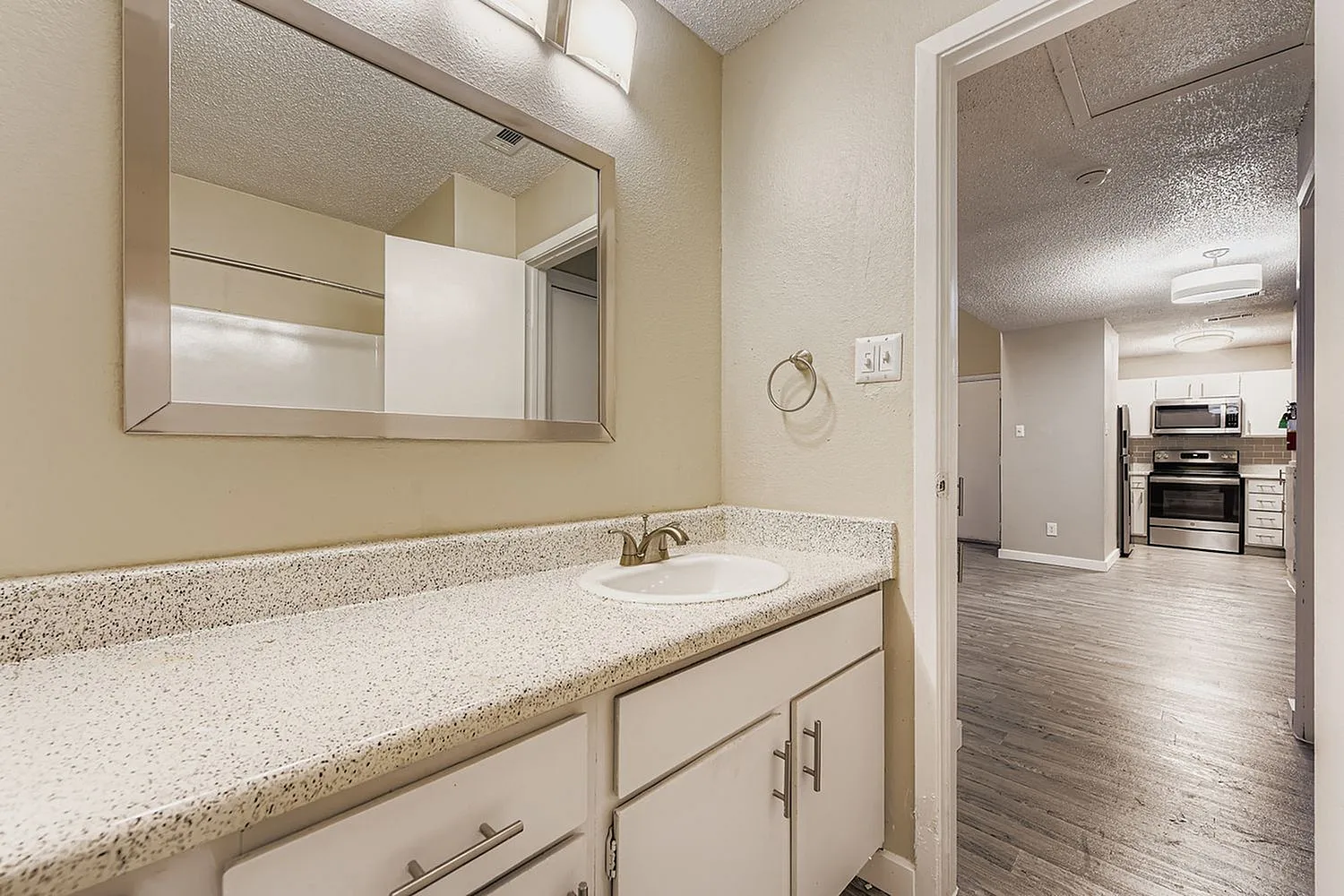 Bathroom, speckled countertops, white cabinetry, rectangular mirror, wood-style plank flooring in adjacent kitchen