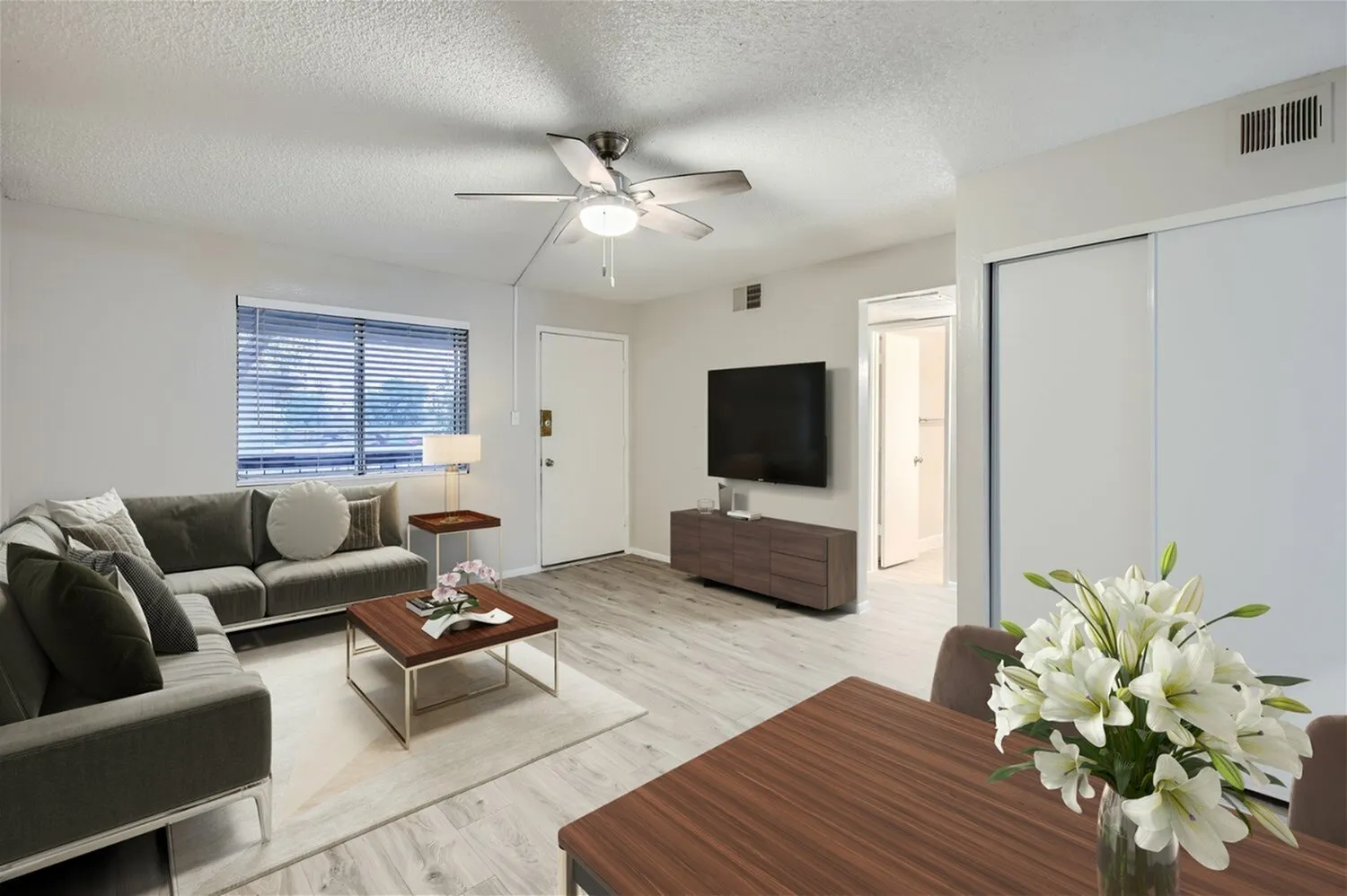 Living room, wood-style plank flooring, gray sofa, ceiling fan, mounted TV, dining table with white flower arrangement