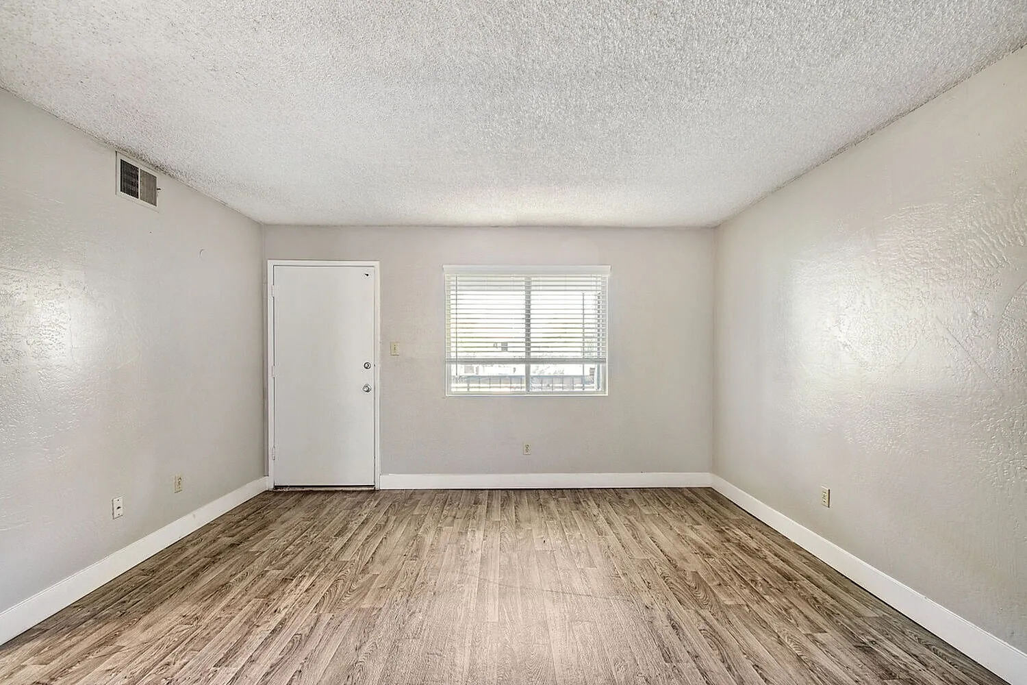Empty room, wood-style plank flooring, white painted walls, window with shutters, entry door