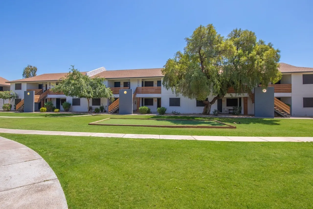 Multi-unit residential buildings, green lawn, mature trees, paved walkway, clear sky, Phoenix AZ