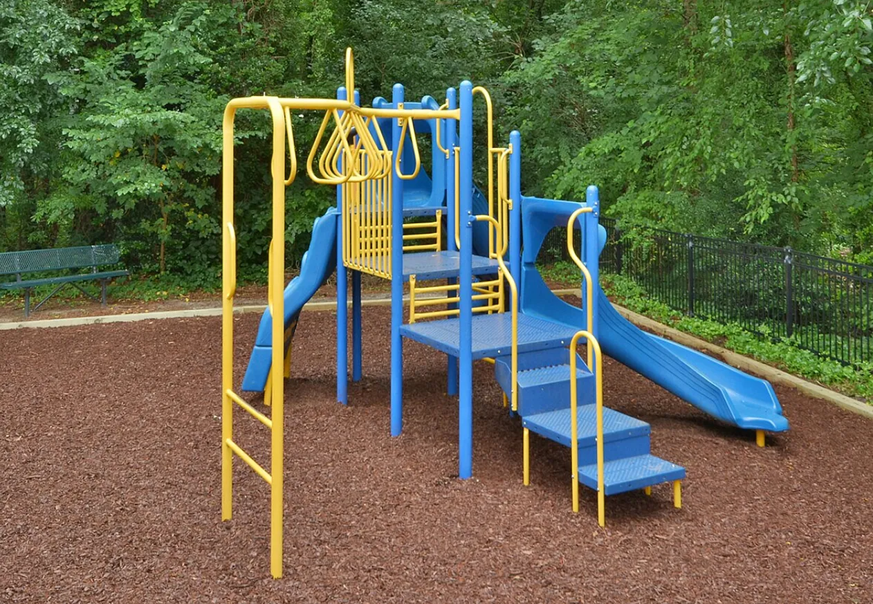 Playground, blue and yellow metal structure, slide, climbing elements, mulch surfacing, wooded backdrop