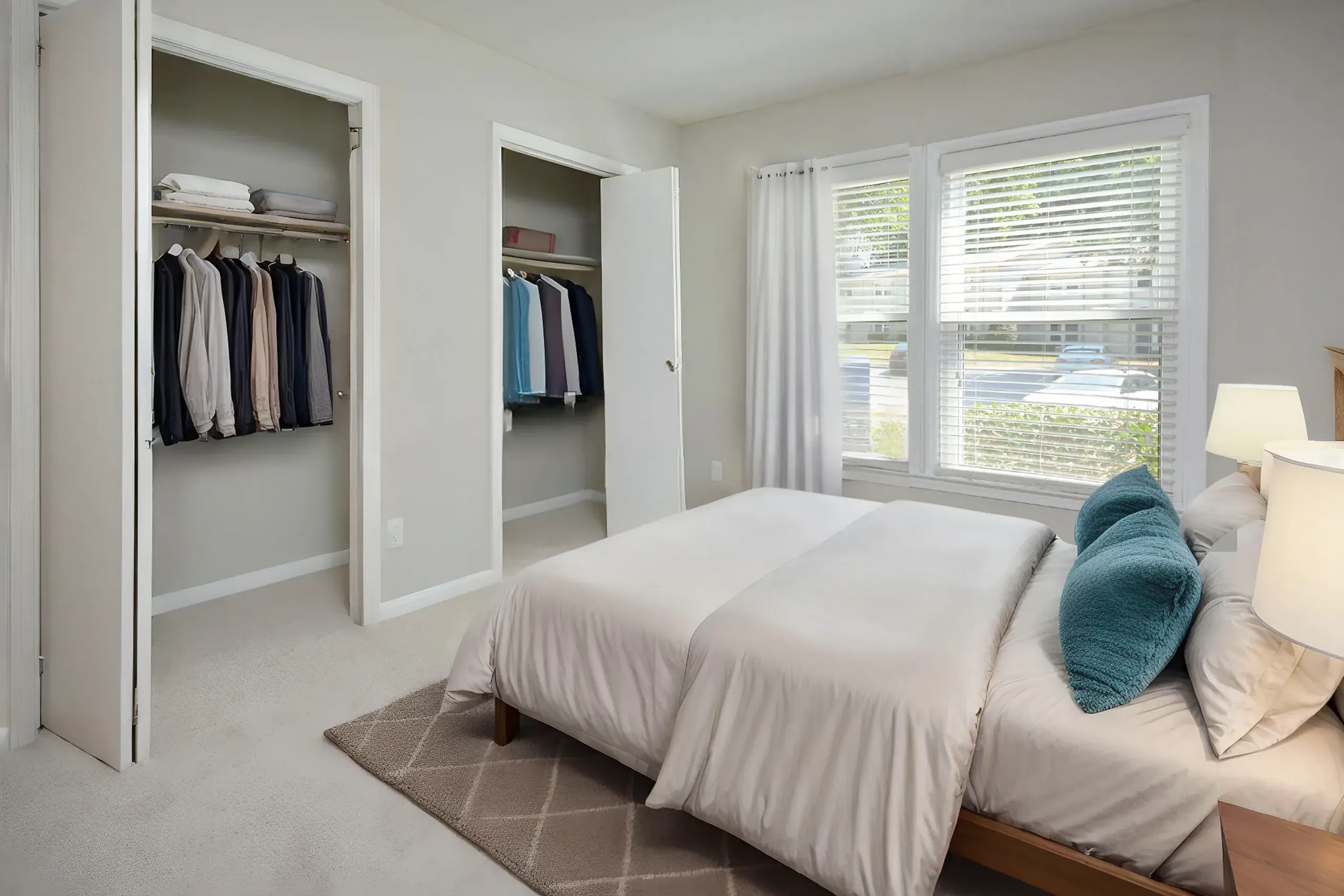 Primary bedroom, dual walk-in closets, glass doors, bed with white bedding, large windows with white curtains, carpet flooring, table lamps