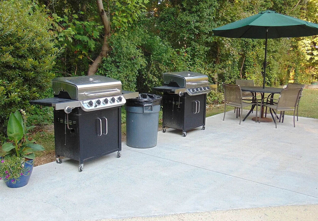 Patio with multiple stainless grills, trash bin, dining table, umbrella, landscaping